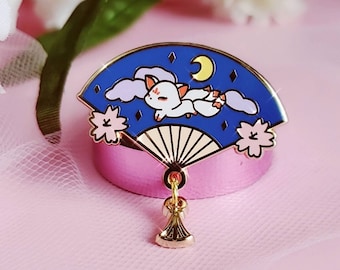 May include: Gold enamel pin featuring a white fox sleeping on a blue and white fan with a crescent moon and stars. The fan has a gold tassel.