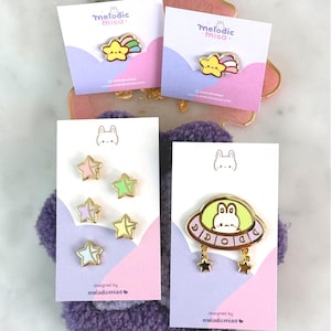 May include: Enamel pins on cards featuring a yellow star with a rainbow, star-shaped pins in pastel colors, and a bunny in a UFO design. The cards have the text "melodic misa" and "designed by melodicmisa".