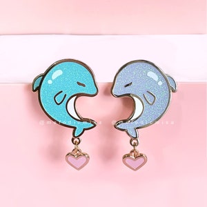 May include: A pair of gold-tone dolphin earrings with a pink heart charm. The dolphins are outlined in black and have a glittery finish. One dolphin is blue and the other is purple.