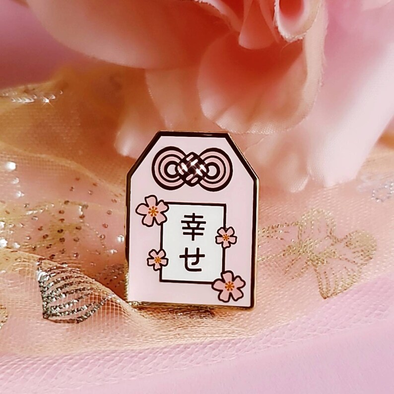 May include: A pink enamel pin with a white square in the center that says "幸せ" (shiawase) which means happiness in Japanese. The pin is decorated with pink cherry blossoms and a gold knot.