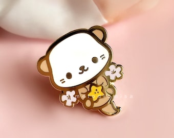May include: Gold-tone enamel pin featuring a white cartoon bear holding a yellow star and white flowers. The bear has a brown nose and black eyes.