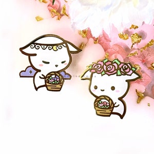 Könnte beinhalten: Zwei goldfarbene Emaille-Pins mit weißen, geisterhaften Figuren mit Blumenkränzen. Ein Geist hält einen Korb mit rosa und lila Blumen, während der andere einen Korb mit rosa Blumen hält. Beide Figuren haben schwarze Augen und rosa Wangen.