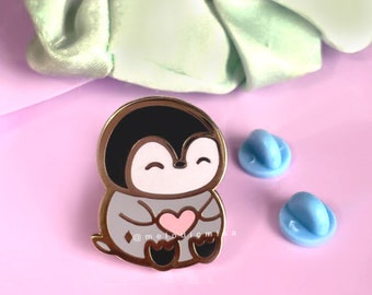 May include: Gold-tone enamel pin featuring a cartoon penguin holding a pink heart. The penguin is wearing a gray sweater and has a black head and white face. The pin is on a purple background.