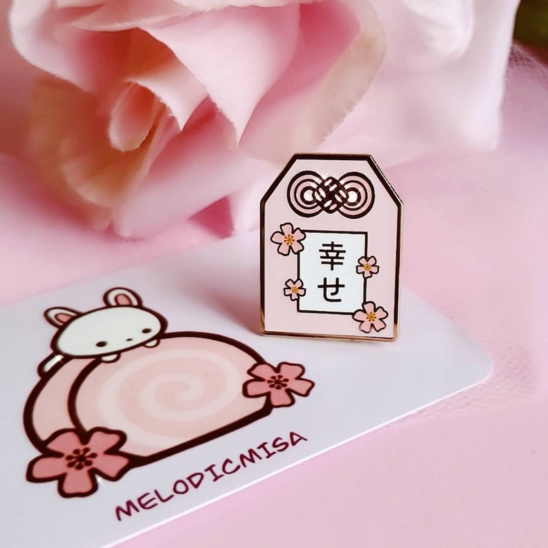 May include: A pink enamel pin with a gold outline. The pin features a Japanese charm with a white rectangle that says "幸せ" (shiawase) which means happiness in Japanese. The charm is decorated with pink cherry blossoms.