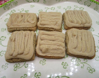 May include: Six square-shaped cookies arranged in two rows on a white plate with a green clover pattern. Each cookie is topped with a thick layer of light brown, piped frosting that features a distinct, continuous swirling texture across the surface.