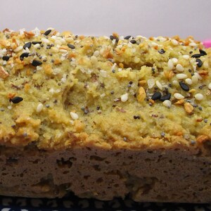 May include: A loaf of bread with a golden-brown crust and a light green interior. The top is generously sprinkled with a mix of seeds, including black sesame, white sesame, and poppy seeds. The bread sits on a blue and white patterned plate.