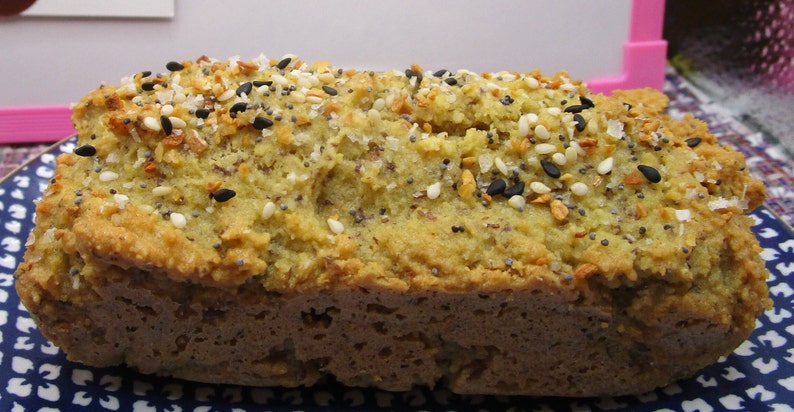 May include: A loaf of bread with a golden-brown crust and a light green interior. The top is generously sprinkled with a mix of seeds, including black sesame, white sesame, and poppy seeds. The bread sits on a blue and white patterned plate.