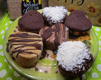 May include: A plate of heart-shaped baked goods, some covered in dark chocolate, others with coconut flakes or chocolate drizzle. The plate sits on a green and white polka dot surface. A sign says "Welcome to our".