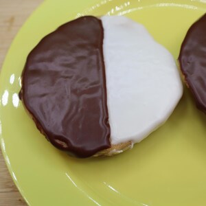 May include: Two black and white cookies on a yellow plate. Each cookie is round with a half-chocolate and half-vanilla frosting. The plate is on a wooden cutting board with a green and white polka dot cloth.