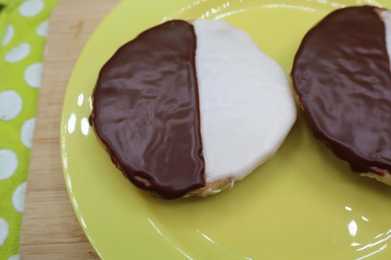 May include: Two black and white cookies on a yellow plate. Each cookie is round with a half-chocolate and half-vanilla frosting. The plate is on a wooden cutting board with a green and white polka dot cloth.