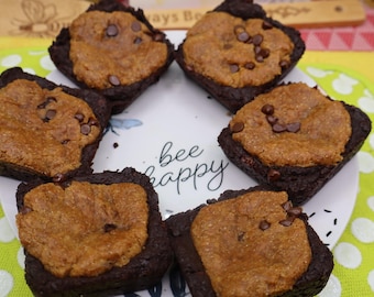 Può includere: Sei dolcetti quadrati ibridi brownie e biscotto disposti in cerchio su un piatto bianco. Ogni quadrato ha una base di brownie al cioccolato fondente sormontata da uno strato di biscotto dorato con gocce di cioccolato. Il piatto riporta la scritta bee happy.