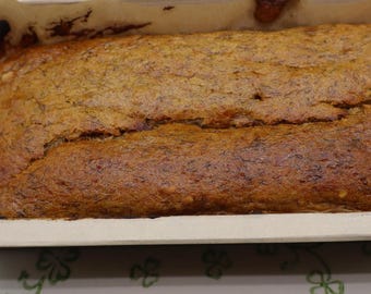 Könnte beinhalten: Ein frisch gebackener Brotlaib in einer rechteckigen Backform aus Papier. Das Brot hat eine goldbraune Kruste mit einer leicht gerissenen Oberseite. Die Form steht auf einem weißen Teller mit grünen Kleemotiven.