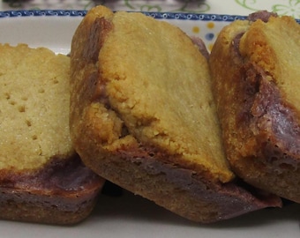 May include: Three square-shaped baked goods arranged in a row on a white oval plate with a blue and yellow patterned rim. Each item features a golden-brown, textured top layer resting on a dense, dark purple base with visible fruit-like coloration.