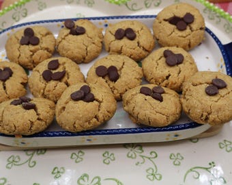 Puede incluir: Una fuente rectangular de cerámica blanca con borde azul y un patrón de tréboles verdes contiene doce galletas redondas de color marrón dorado. Cada galleta tiene una textura agrietada y está cubierta con dos o tres chispas de chocolate.