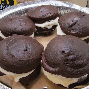 May include: Five chocolate whoopie pies with white frosting filling, arranged in a silver baking pan lined with parchment paper.