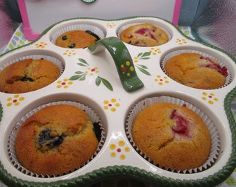 May include: A white ceramic muffin pan with five circular compartments, each holding a baked muffin. The pan features a green central handle and a decorative pattern of small yellow and red flowers. The edges are scalloped with a green textured rim.