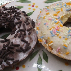 May include: Two round donuts on a patterned plate. The left donut has chocolate icing with dark brown chocolate sprinkles. The right donut has a light-colored glaze topped with small, multi-colored sprinkles in shades of pink, blue, and orange.