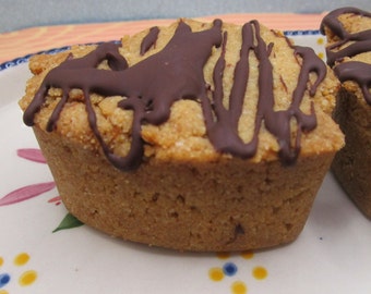 May include: A golden-brown, heart-shaped baked good with a textured, crumbly surface drizzled with dark chocolate. The treat has a dense, rounded edge and sits on a white plate decorated with small, colorful floral patterns in pink, yellow, and blue.