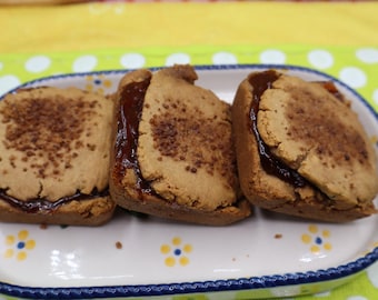 May include: Three square-shaped sandwich biscuits with a light brown, textured surface and a dark brown jam filling, arranged in a row on a white rectangular ceramic tray featuring a blue rim and small yellow and blue floral patterns.