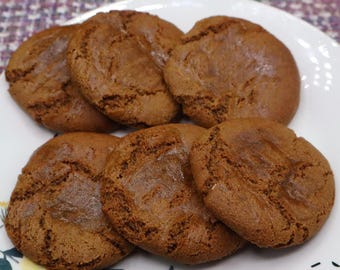 May include: Six round, brown, baked cookies with a textured, cracked surface arranged on a white plate. The cookies have a uniform dark brown color and a slightly uneven, rustic appearance with visible ridges and small fissures across their tops.