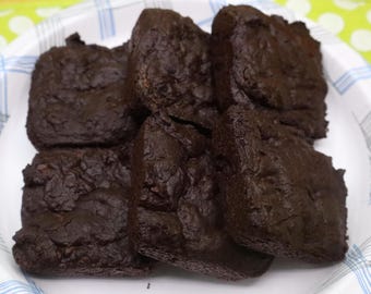 Puede incluir: Cinco brownies de chocolate negro de forma cuadrada con una superficie densa y texturizada, dispuestos sobre un plato blanco. Los brownies tienen un color marrón intenso y un aspecto horneado ligeramente irregular con grietas visibles.
