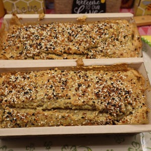 May include: Two loaves of bread in wooden baking molds, topped with a mix of seeds and spices. The bread is light brown, and the seeds are black, white, and tan. The loaves sit on a decorative plate with a green and white border.