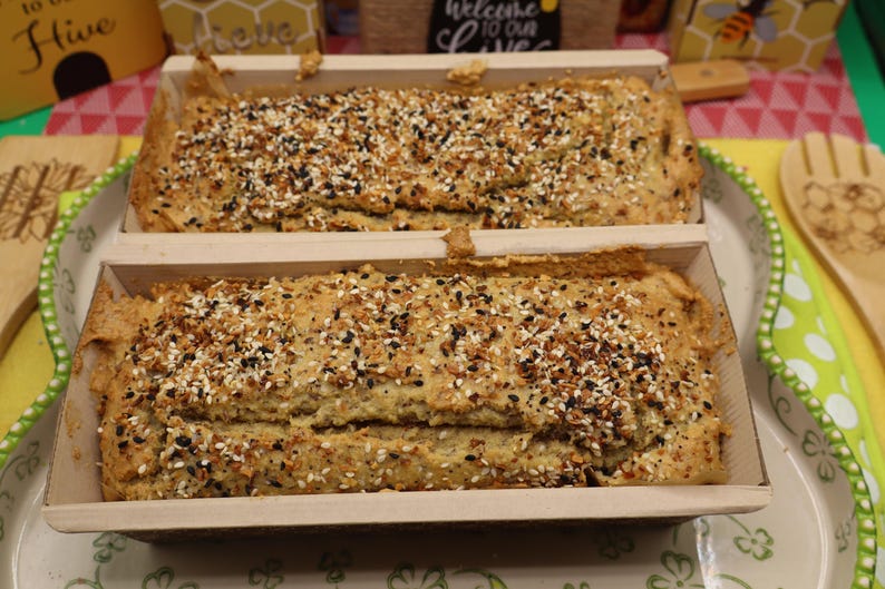 May include: Two loaves of bread in wooden baking molds, topped with a mix of seeds and spices. The bread is light brown, and the seeds are black, white, and tan. The loaves sit on a decorative plate with a green and white border.