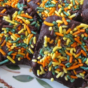 Puede incluir: Primer plano de trozos de corteza de chocolate cubiertos de chispas de colores. La corteza es de color marr&oacute;n oscuro y las chispas son amarillas, naranjas, verdes y marrones. Las piezas est&aacute;n dispuestas sobre un plato decorativo.