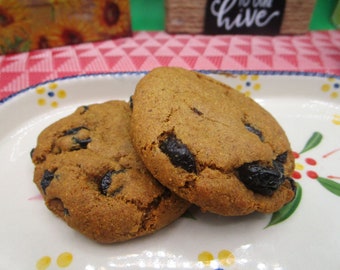 Peut inclure: Deux biscuits ronds brun doré avec des raisins secs foncés visibles incrustés dans la pâte, reposant sur une assiette ovale en céramique blanche décorée de petits points jaunes et de motifs de feuilles vertes sur le bord festonné bleu.