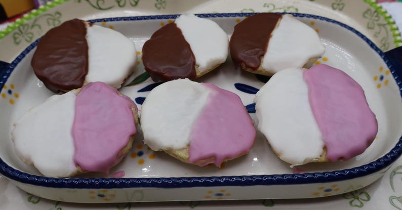 May include: Six black and white cookies on a decorative oval plate. Three cookies have a white and chocolate frosting, and three have white and pink frosting. The plate has a blue and white floral design.
