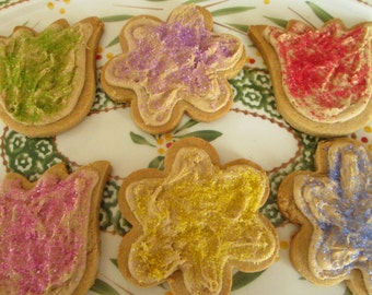 May include: Six flower-shaped and tulip-shaped cookies with colorful frosting and sprinkles. The cookies are decorated with green, purple, red, pink, yellow, and blue sprinkles. The cookies are on a decorative plate.