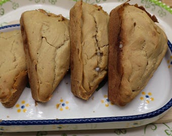 Puede incluir: Cuatro pasteles horneados de color dorado, posiblemente bollos o galletas, dispuestos en una fuente ovalada decorativa. Los pasteles tienen una superficie ligeramente agrietada y se presentan en una fuente blanca con detalles florales azules y amarillos y un asa azul.