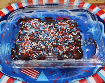 May include: A rectangular chocolate-covered treat topped with red, white, and blue sprinkles, housed in a clear plastic container. The treat sits on a round paper plate featuring a blue background with red and white firework and flag patterns.