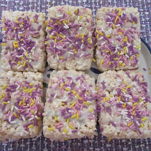 May include: Six rectangular rice crispy treats with white frosting and colorful sprinkles. The treats are arranged on a decorative plate with a blue rim and floral accents. The sprinkles are a mix of purple, pink, yellow, and white.