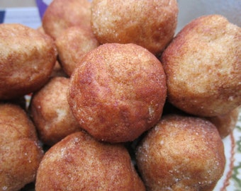 May include: A close-up shot of a pile of cinnamon sugar-coated donut holes. The golden-brown spheres are dusted with cinnamon and sugar, creating a textured surface. The image highlights the freshly baked appearance of the treats.