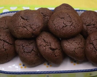 Puede incluir: Una colección de galletas de chocolate de color marrón oscuro, redondas y con superficies agrietadas, apiladas dentro de una fuente ovalada de cerámica blanca. El plato tiene asas azul oscuro y un patrón decorativo azul y amarillo.