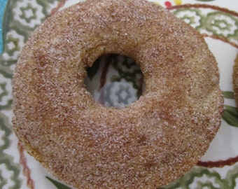 Könnte beinhalten: Ein runder, goldbrauner Back-Donut, der mit einer feinen Schicht aus granuliertem Zucker und Zimt überzogen ist. Der Donut hat ein Loch in der Mitte und eine texturierte, körnige Oberfläche, auf der die hellbraune Gewürzmischung gleichmäßig verteilt ist.