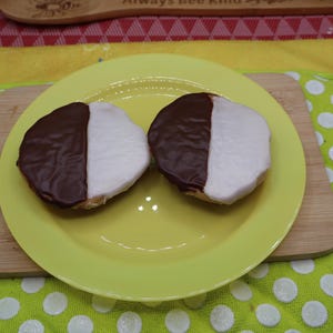 May include: Two black and white cookies, each with a half-chocolate and half-vanilla frosting, sit on a yellow plate. The plate rests on a wooden cutting board. The background includes a yellow cloth and a green potholder with white polka dots.
