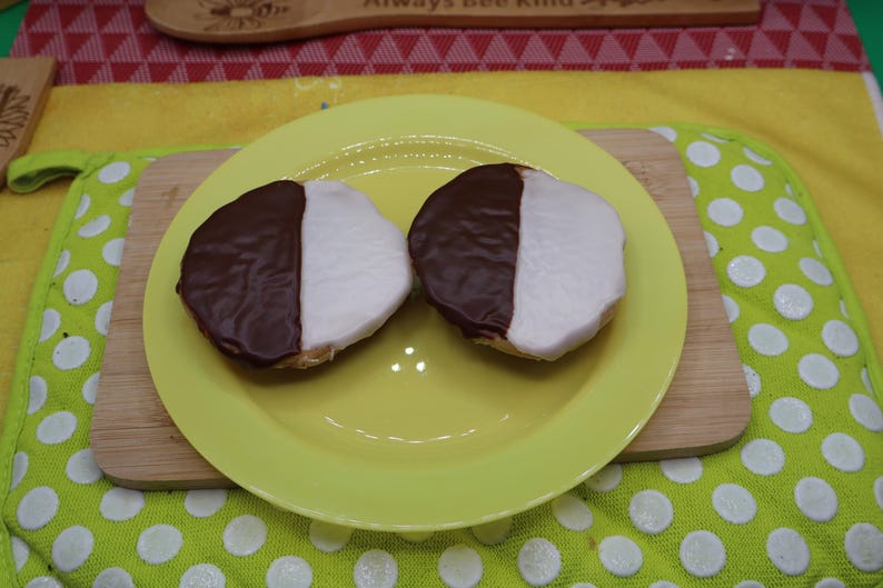 May include: Two black and white cookies, each with a half-chocolate and half-vanilla frosting, sit on a yellow plate. The plate rests on a wooden cutting board. The background includes a yellow cloth and a green potholder with white polka dots.