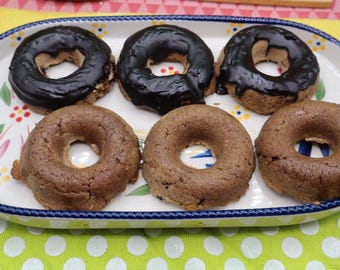 May include: Six ring-shaped baked goods arranged on an oval ceramic platter with a blue rim and a floral pattern. Three of the items are topped with a smooth, dark chocolate glaze, while the other three remain plain, showing a light brown texture.