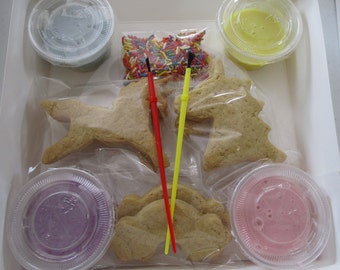 May include: A biscuit decorating kit containing several plain unicorn and rainbow-shaped sugar biscuits, four small plastic containers of coloured icing, a small bag of multi-coloured sprinkles, and two thin plastic decorating tools in red and yellow.