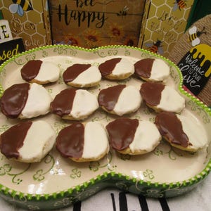 May include: A decorative, oval-shaped platter holds a dozen black and white cookies. Each cookie is half-covered in white icing and half in dark chocolate. The platter has a green scalloped edge and a floral design.