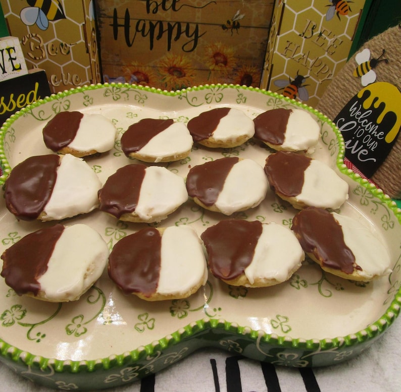 May include: A decorative, oval-shaped platter holds a dozen black and white cookies. Each cookie is half-covered in white icing and half in dark chocolate. The platter has a green scalloped edge and a floral design.