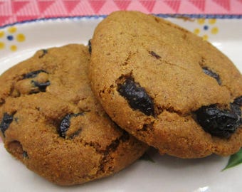 May include: Two round, golden-brown baked cookies with a textured surface and visible dark, dried fruit pieces embedded throughout. The cookies are positioned on a white ceramic plate featuring a decorative pattern of small blue and yellow dots.