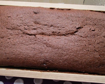 Op de afbeelding: Een rechthoekige chocoladecake gebakken in een lichtgekleurde houten vorm. De cake heeft een donkerbruin, getextureerd en licht gebarsten oppervlak dat de gehele lengte en breedte van de houten bak vult, met een gebakken uiterlijk.
