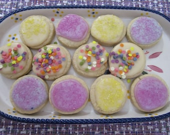 Puede incluir: Una bandeja rectangular de cerámica con borde festoneado azul y detalles florales contiene doce galletas redondas. Las galletas están cubiertas con glaseado blanco, rosa o amarillo, algunas decoradas con chispas de colores o azúcar brillante.