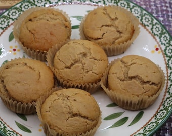 May include: Six golden-brown muffins with cracked tops, each contained in a light brown paper liner, arranged on a white ceramic plate featuring a decorative green and red floral pattern along the scalloped rim and a purple textured surface.