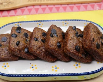 Könnte beinhalten: Sechs rechteckige, braune Bananenbrotscheiben mit dunklen Schokoladenstückchen, die in einer Reihe auf einer ovalen, weißen Keramik-Servierplatte mit blauem Rand und kleinen gelb-blauen Blumenmustern am Innenrand angeordnet sind.