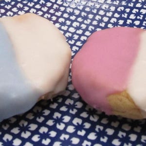 May include: Two frosted cakes, one blue and white, the other pink and white, sit on a blue and white patterned plate. The cakes are oval-shaped and have a smooth, glossy frosting. The plate has a floral design.