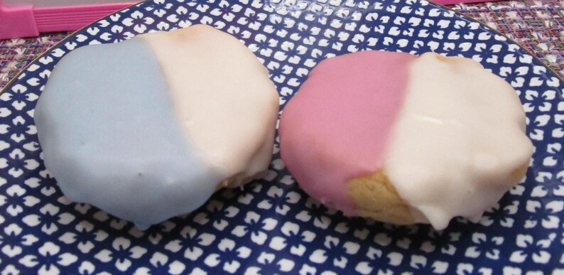 May include: Two frosted cakes, one blue and white, the other pink and white, sit on a blue and white patterned plate. The cakes are oval-shaped and have a smooth, glossy frosting. The plate has a floral design.